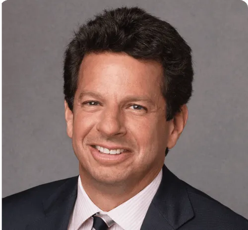 Smiling man with dark curly hair wearing a dark suit jacket, light-colored dress shirt, and striped tie against a gray background.