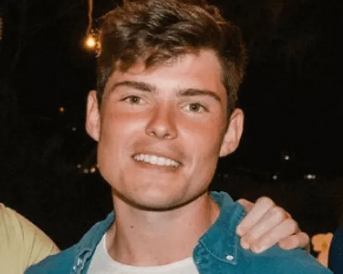 Smiling young man with short brown hair wearing a blue shirt over a white t-shirt at night.