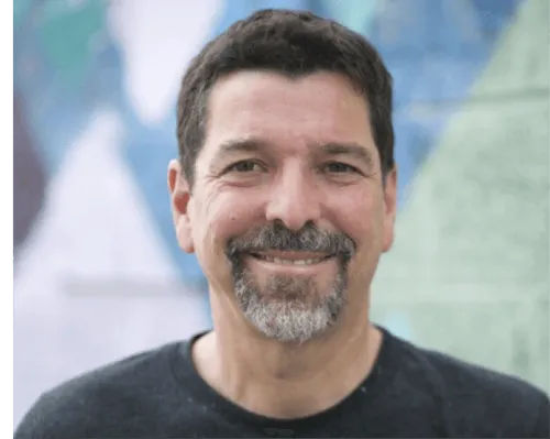 Smiling man with dark hair and a salt-and-pepper beard wearing a dark shirt, standing in front of a blurry colorful background.