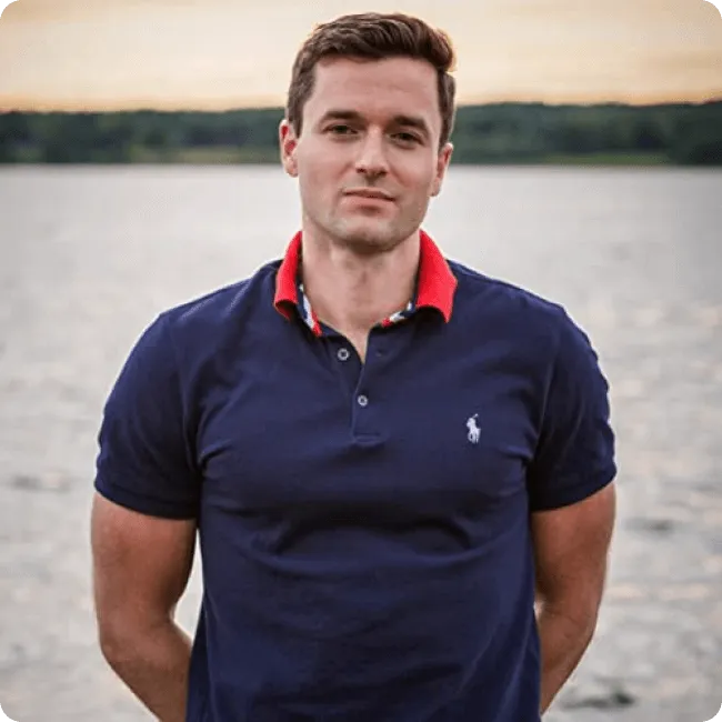 Man in a navy blue polo shirt with a red collar standing in front of a body of water during sunset.