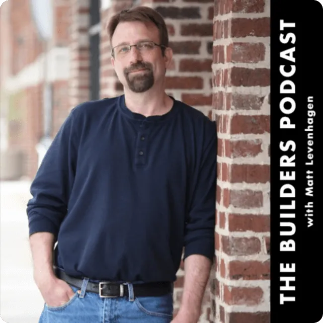 Man with glasses and beard leaning against a brick wall, wearing a navy blue shirt and jeans.