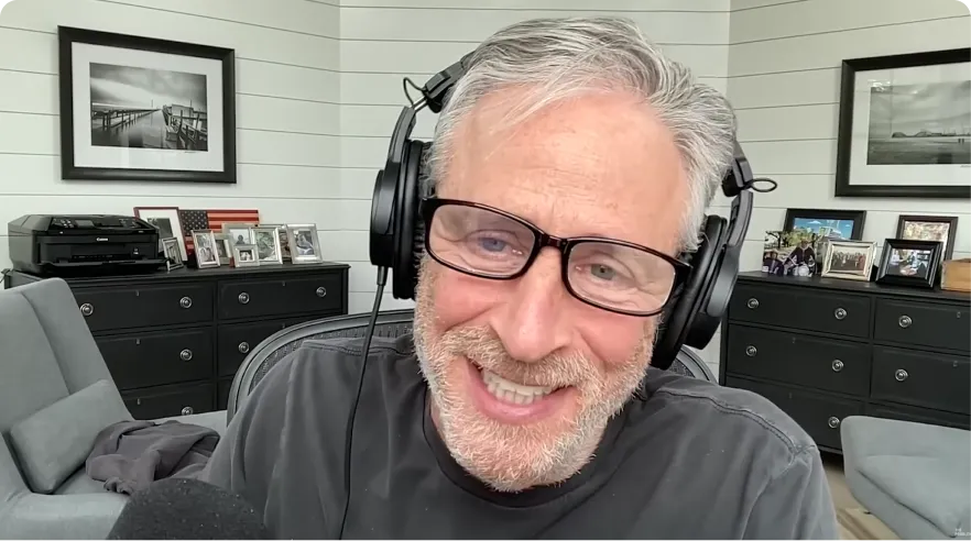 Smiling man with gray hair, beard, and glasses wearing black headphones in a bright room with framed photos and furniture.