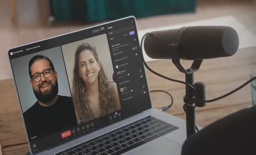 Laptop screen showing a video call with a smiling man and woman, and a microphone on a wooden table nearby.