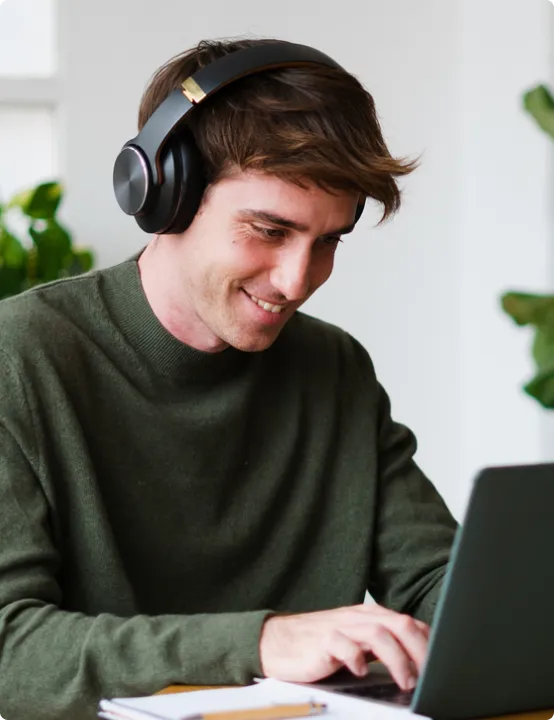 Smiling man wearing black headphones typing on a laptop at a desk with plants in the background.
