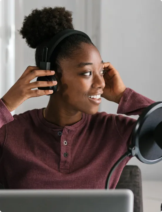 Smiling woman with curly hair and headphones sitting at a desk with a laptop and a microphone.