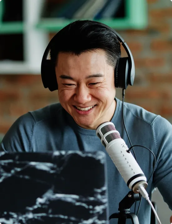 Smiling man wearing headphones speaking into a microphone in front of a laptop with a black marble cover.