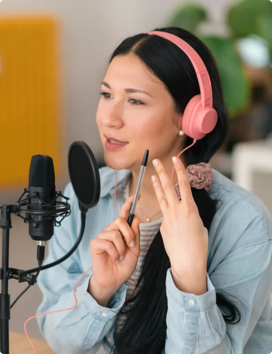 Woman with long black hair wearing pink headphones and holding a pen, speaking into a microphone with a pop filter.