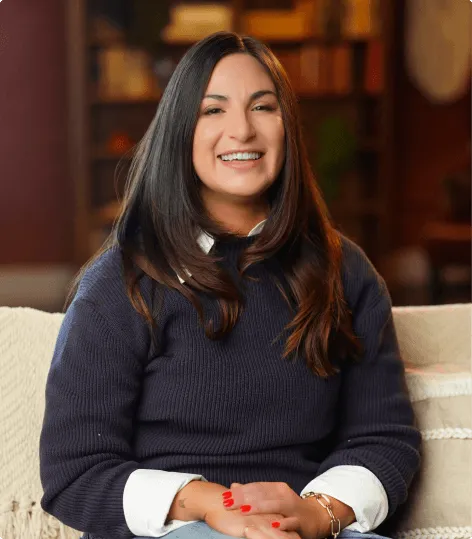 Smiling woman with long dark hair wearing a navy blue sweater and white shirt sitting on a couch.