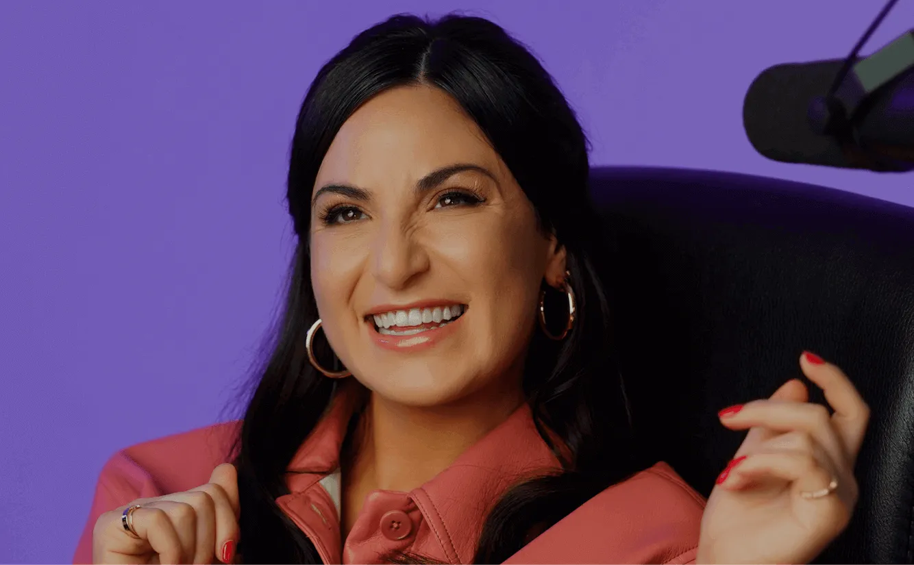 Smiling woman with long dark hair wearing hoop earrings and a pink jacket against a purple background.