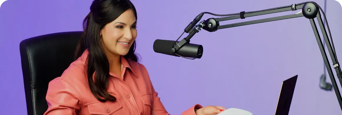 Woman in a pink jacket sitting at a desk speaking into a microphone with a laptop in front of her.