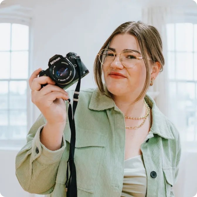 Young woman with glasses and layered necklaces holding a Nikon camera and smiling indoors.
