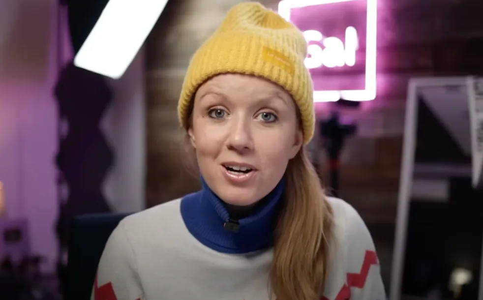 Woman wearing a yellow beanie and a blue turtleneck sweater speaking in a softly lit room with a neon sign in the background.