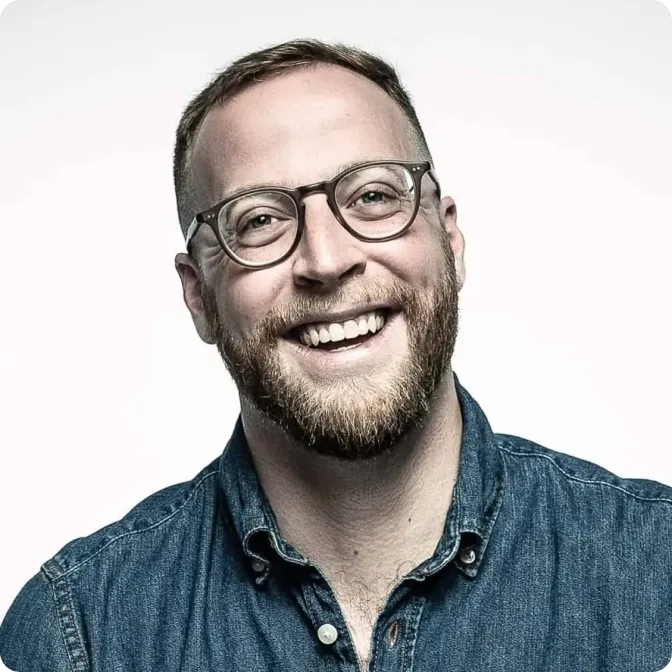 Smiling man with beard and glasses wearing a blue denim shirt against a white background.