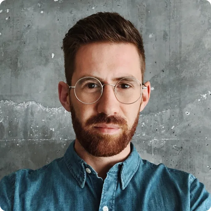 Portrait of a man with brown hair, beard, round glasses, and a blue button-up shirt against a gray concrete wall.
