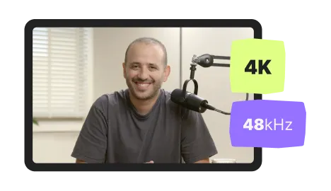 Smiling man sitting in front of a microphone in a well-lit room with window blinds.