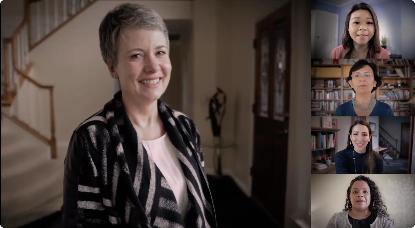 Smiling woman standing in a warmly lit home, with four other women appearing on a video call screen to the right.