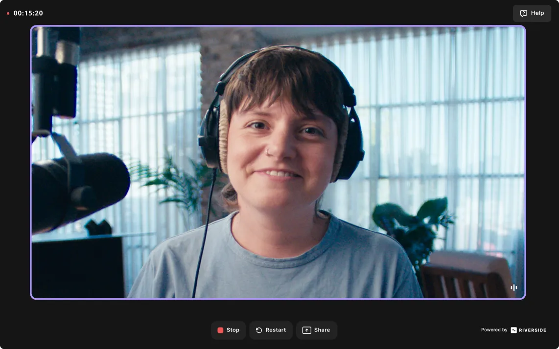 Smiling person wearing headphones and a light blue shirt, sitting in front of a microphone in a room with plants and large windows.
