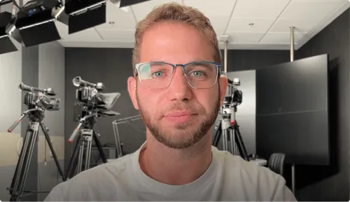 Man with short hair and glasses standing in a studio with professional cameras on tripods.
