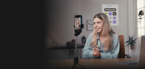 Young woman with long blonde hair speaking during a video call recorded on a smartphone mounted on a tripod, sitting in a bright room with podcast posters on the wall.