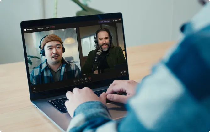 Person typing on a laptop during a video call showing two men wearing headphones and speaking.