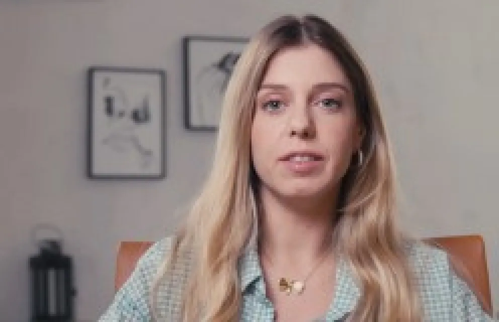 Young woman with long blonde hair wearing a light blue checkered shirt sitting indoors with framed abstract art on the wall behind her.