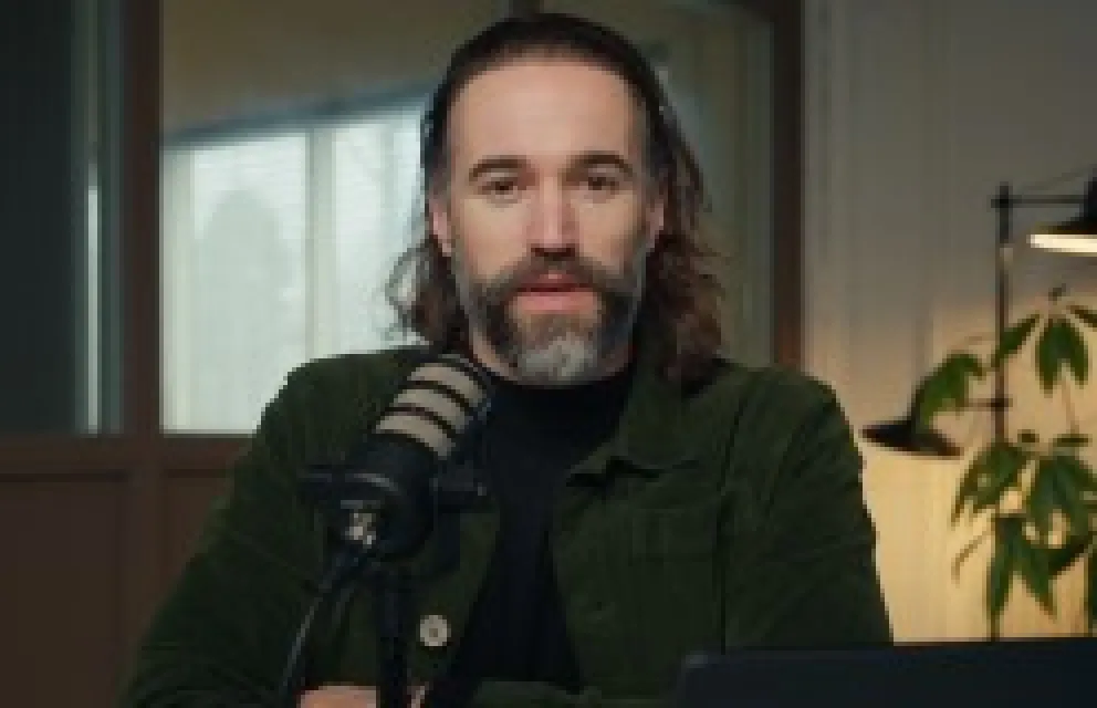 Man with medium-length hair and a beard speaking into a microphone in a cozy room with a window and plant in the background.