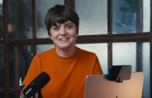 Person with short dark hair smiling, wearing an orange shirt, sitting at a desk with a microphone and a laptop.