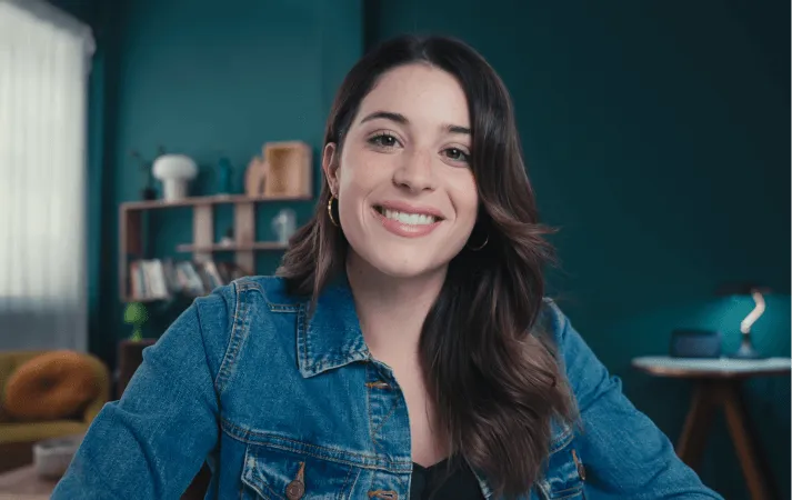 Smiling woman with long dark hair wearing a denim jacket, sitting indoors in a cozy room with shelves and a desk lamp in the background.