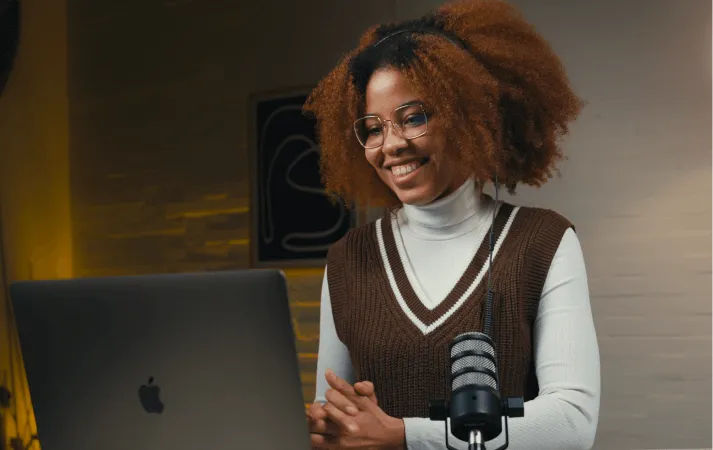 Smiling woman with curly hair and glasses speaking in front of a laptop and microphone.