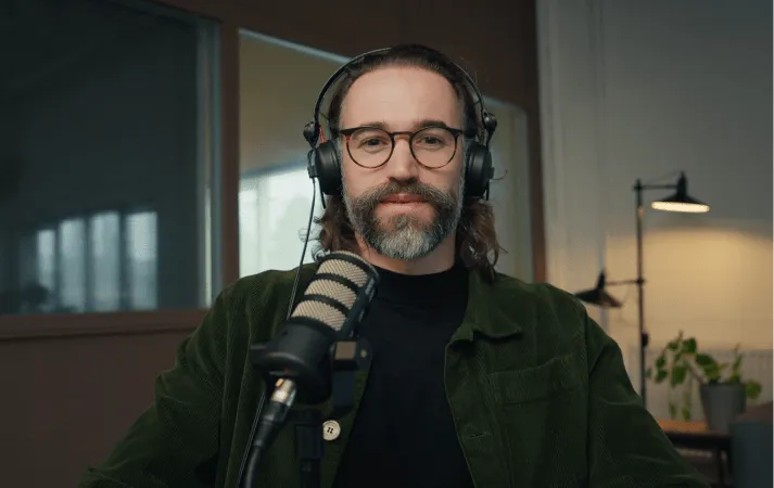 Man with glasses and beard wearing headphones speaking into a microphone in a recording studio.