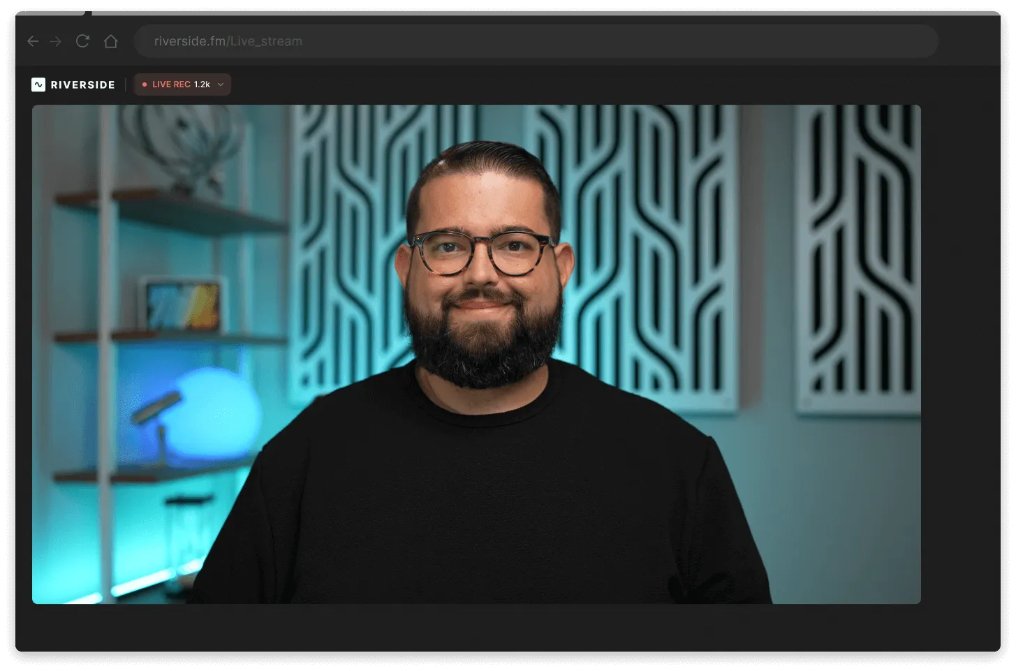 Smiling man with glasses and a beard in a black shirt sitting in a well-lit room with patterned wall panels and a glowing blue lamp on shelves behind him.