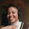 Smiling young woman with curly hair wearing headphones and a brown sweater against a dark background.