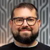 Smiling man with glasses, beard, and mustache wearing a black shirt in front of a patterned background.