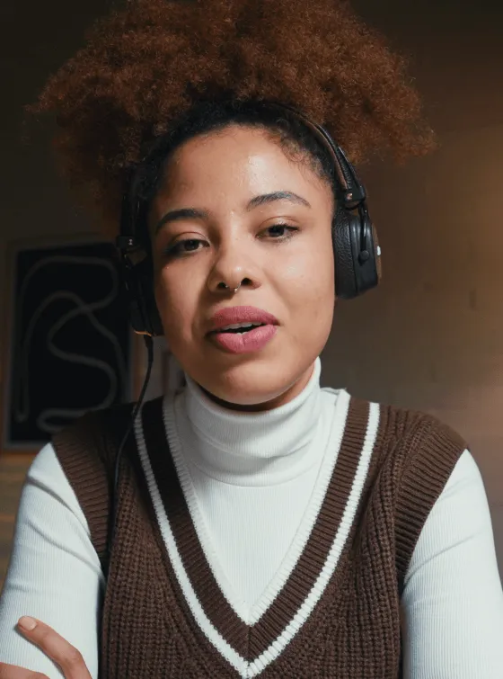 Young woman with curly hair wearing a brown sweater vest and white turtleneck, speaking while wearing black headphones.