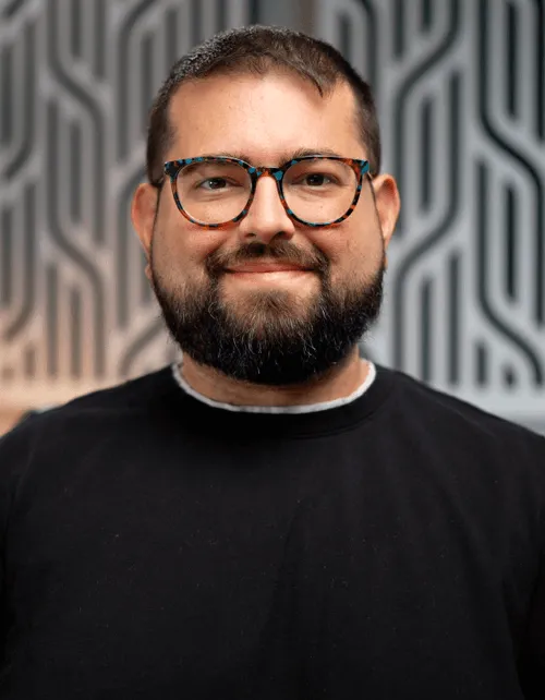 Smiling man with beard and tortoiseshell glasses wearing a black shirt in front of a black and white patterned background.
