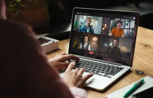 Person typing on a laptop participating in an online video meeting with four colleagues visible on the screen.