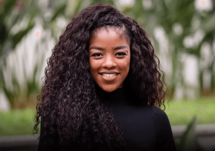 Smiling woman with long curly hair wearing a black turtleneck sweater standing outdoors with blurred greenery background.