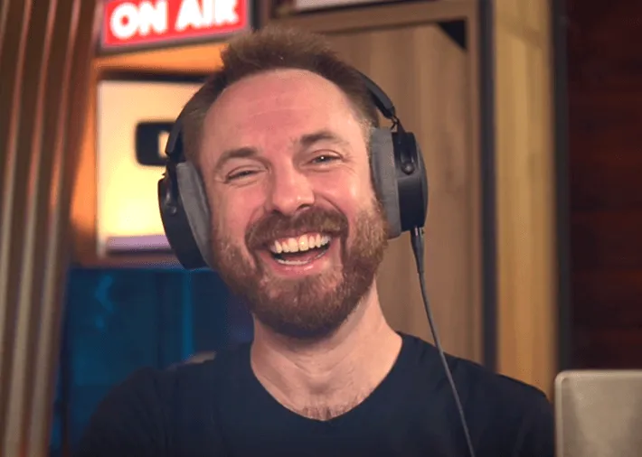 Smiling man with beard wearing large headphones in front of an 'ON AIR' sign in a recording studio.