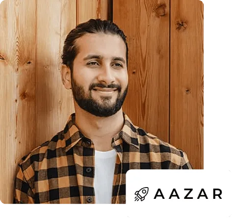 Smiling man with beard and tied-back hair wearing a checkered shirt standing against a wooden wall.
