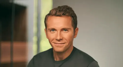 Smiling man with short light brown hair wearing a dark shirt, with a blurred background.