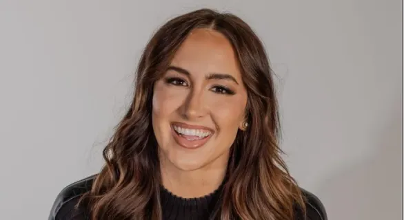 Smiling woman with long brown hair wearing a black top against a plain light background.