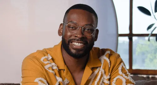 Smiling man with glasses and beard wearing a mustard yellow shirt with white patterns.
