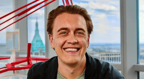 Smiling man with light brown hair in a black shirt sitting indoors near large windows with a city view and red art installation.