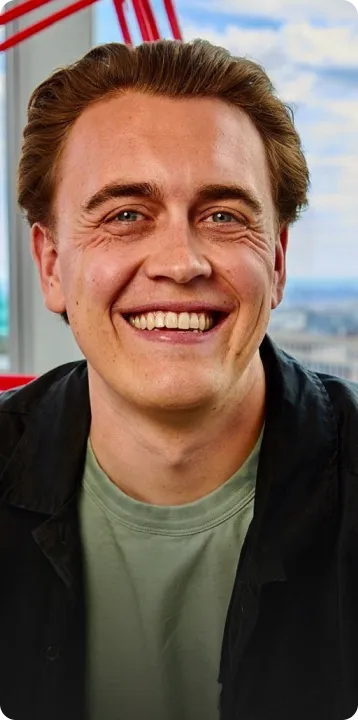 Smiling man with light brown hair wearing a green shirt and dark jacket, with a cityscape visible through a window behind him.