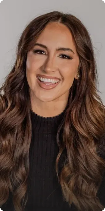 Woman with long wavy brown hair smiling and wearing a black top against a gray background.