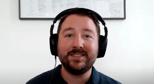 Man with beard wearing black headphones smiling in front of a white wall with framed documents.