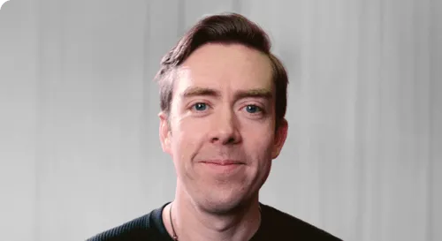 Portrait of a smiling man with light skin and brown hair wearing a black shirt against a gray background.