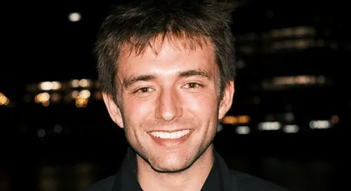 Smiling young man with short brown hair against a dark background with blurred lights.
