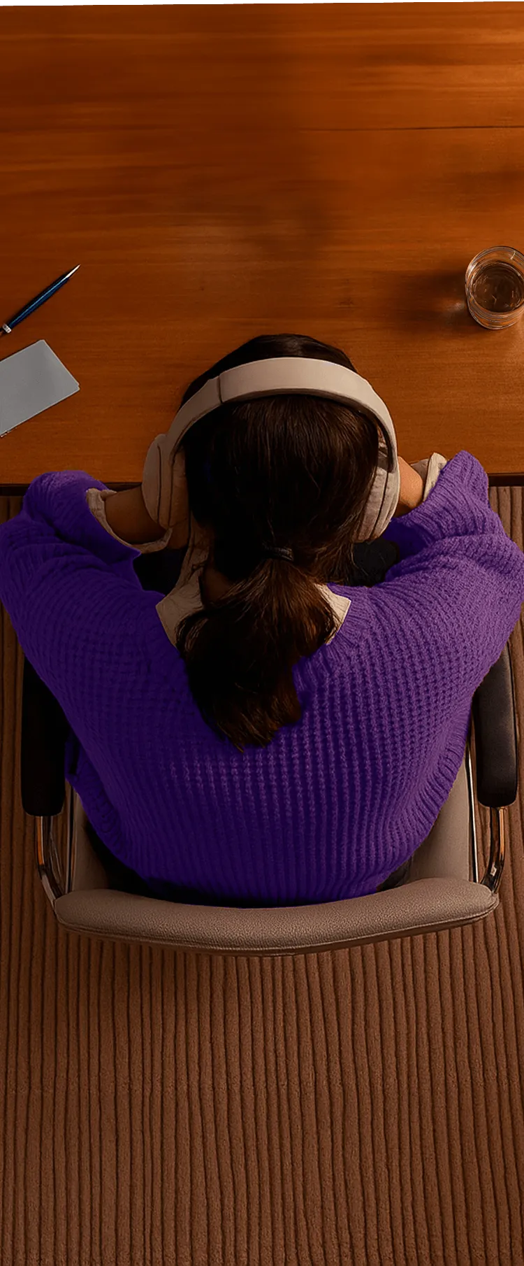 Person wearing beige headphones and a purple sweater sitting at a wooden desk with a pen, notepad, and glass of water.