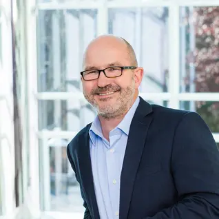 Smiling professional in glasses and navy blazer standing near windows